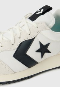 Baskets blanches avec des accents noirs, présentant une tige en toile et des détails en daim. Comprend un logo en forme d'étoile noire et une semelle en caoutchouc texturée.