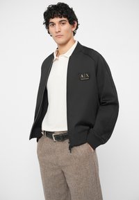 Άνδρας φορώντας μαύρο τζάκετ bomber με το λογότυπο της Armani Exchange, λευκό πόλο μπλουζάκι, καφέ υφασμάτινο παντελόνι και μαύρη ζώνη, με τα χέρια στις τσέπες.