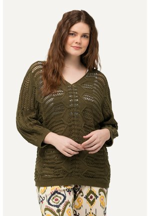 Oliwkowy sweter z dzianiny z dekoltem w serek, o teksturowanym wzorze i luźnym kroju. Zestawiony z wzorzystymi, jasnokolorowymi spodniami w geometryczne wzory.
