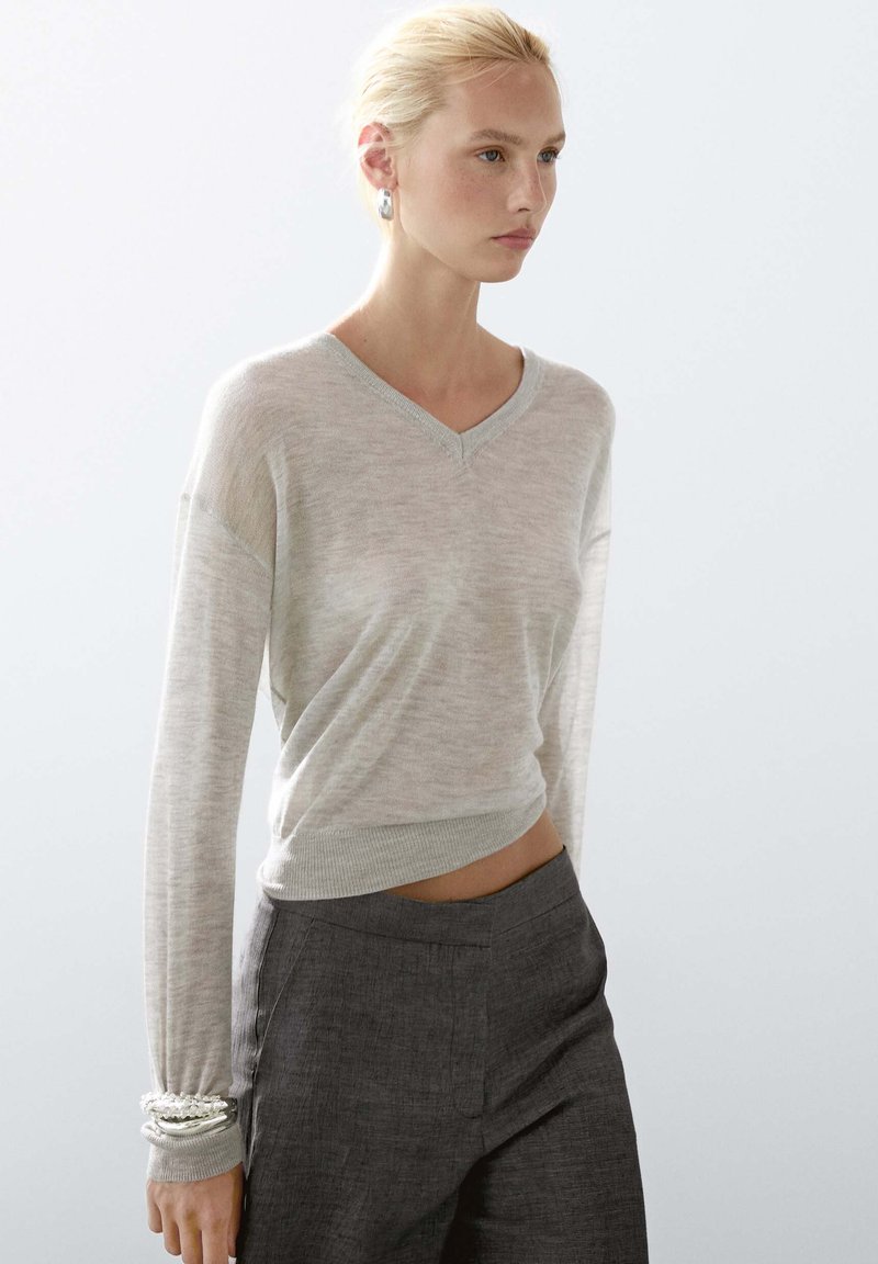 Lichtgrijze V-hals trui met lange mouwen, gebreide textuur en een iets cropped pasvorm, gecombineerd met donkergrijze high-waisted broek.
