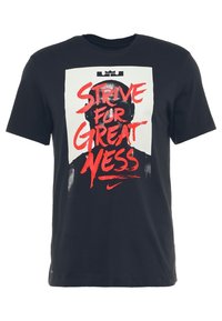 Camiseta de algodón negra con un rectángulo blanco que presenta un texto gráfico en rojo: "ESFUÉRZATE POR LA GRANDEZA" sobre una silueta. Logo de Nike en rojo en la parte inferior.