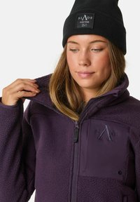 Lila Fleecejacke mit einem Reißverschluss vorne, einer strukturierten Oberfläche und einem Logopatch auf der Brust. Getragen mit einer schwarzen Mütze.