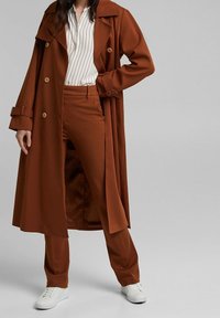 Hnědý trenčkot s velkými knoflíky, odpovídající hnědé kalhoty a bílé pruhované tričko; stylizováno s bílými teniskami. Hladká textura a strukturovaný design.