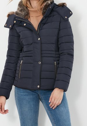 Femme portant une veste matelassée bleu marine avec une capuche doublée de fourrure, un col roulé beige et un jean bleu, debout devant un fond uni.