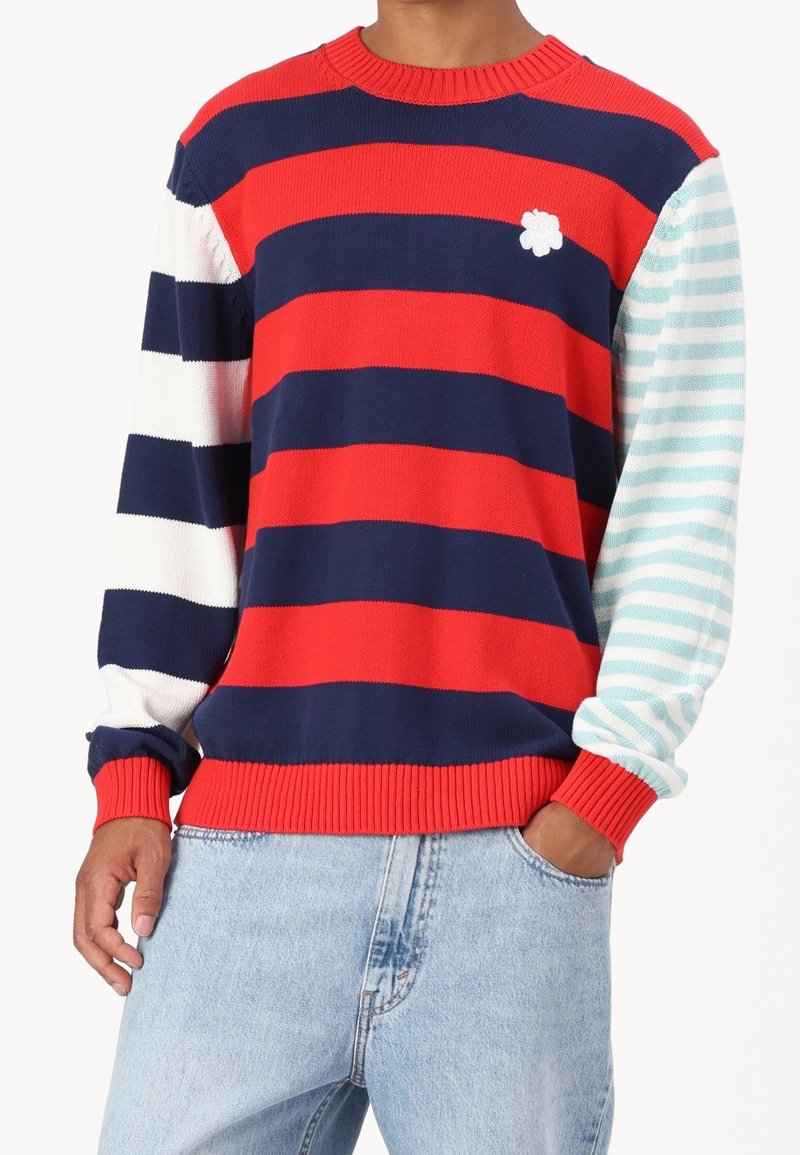 Pull rayé présentant des bandes rouges, bleu marine et blanches avec une manche rayée bleu clair. Encolure et poignets côtelés ; petit accent de fleur blanche.