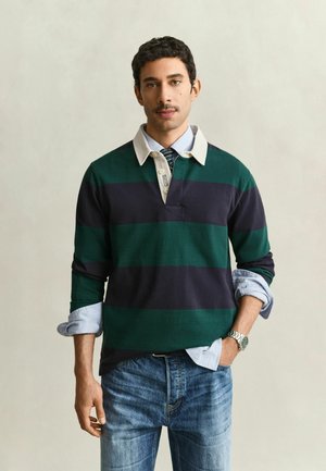 Uomo con capelli ricci che indossa una maglia da rugby a righe verdi e blu scuro sopra una camicia azzurra con le maniche arrotolate e jeans blu.