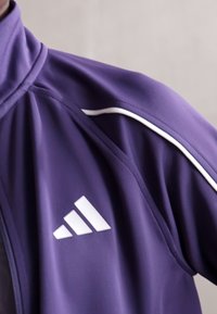 Violet sportinė striukė su baltomis detalėmis, aukštu apykakle ir ryškiu trimis juostelėmis pažymėtu logotipu kairiajame krūtinės kampe. Lygus, lengvas audinys.