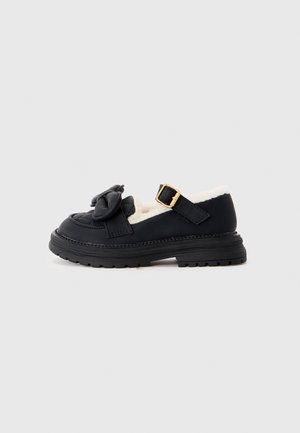 Mocassin en cuir noir avec semelle épaisse, bride à boucle dorée, grand nœud sur le dessus et doublure blanche en peluche visible à l'intérieur.