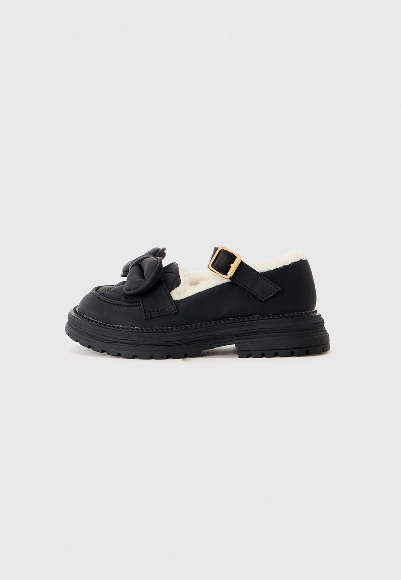 Mocassin en cuir noir avec semelle épaisse, bride à boucle dorée, grand nœud sur le dessus et doublure blanche en peluche visible à l'intérieur.