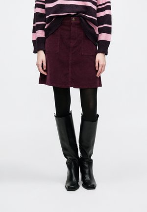 Frau trägt einen lila Cord-Rock, schwarze Strumpfhosen, kniehohe schwarze Lederstiefel und einen schwarz-pink gestreiften Pullover.