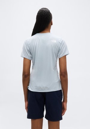 Person med langt fletning set bagfra, iført en lysegrå t-shirt med korte ærmer og mørkeblå shorts mod en ensfarvet baggrund.