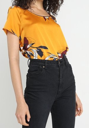Femme portant un haut jaune à fleurs à manches courtes, rentré dans un jean noir taille haute, se tenant devant un fond uni.