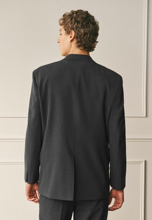 Mann mit lockigem Haar, der eine dunkelgraue Anzugjacke und passende Hose trägt, steht vor einer beigen Wand mit dekorativem Stuck.