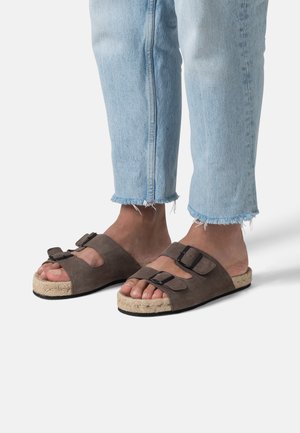 Füße in hellblauen fransigen Jeans mit braunen Wildleder-Sandalen mit doppeltem Schnallenverschluss und gewebten Espadrille-Sohlen vor einem weißen Hintergrund.