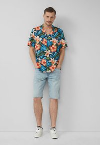 Camicia a maniche corte in stile hawaiano con vivaci motivi floreali e fogliari; abbinata a pantaloni corti in denim blu chiaro e scarpe da ginnastica bianche.
