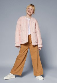 Hele roosa tikitud jakk pikkade varrukatega, mille juurde on sobitatud laiad beežid püksid ja valged spordijalatsid. Lihtne disain, nähtavaid logosid pole.