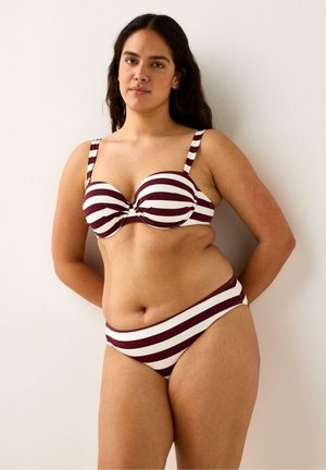 Pitkähiuksinen nainen, jolla on päällään viininpunavalkoraidallinen bikini, seisoo selkä valkoista seinää vasten käsivarret selän takana.