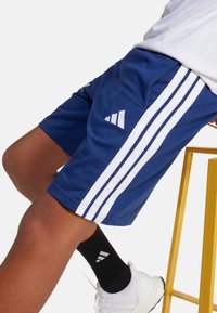 Pantalones cortos deportivos azules con franjas laterales y logo blancos. Diseñados con bolsillos. Usados con calcetas negras y zapatillas blancas sobre un taburete amarillo.