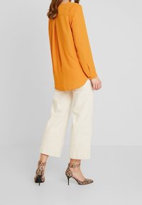 Blusa arancione a maniche lunghe con orlo curvo, abbinata a pantaloni larghi colore avorio. La modella indossa tacchi alti con stampa pitone marrone.