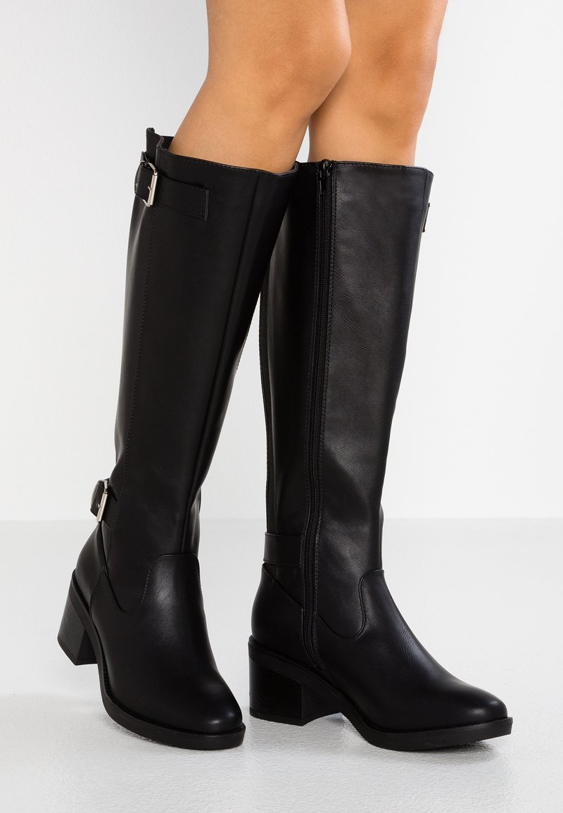 Czarne skórzane kozaki do kolan na słupkowym obcasie, z bocznymi zamkami błyskawicznymi i dekoracyjnymi klamrami, noszone przez osobę stojącą na białej podłodze.