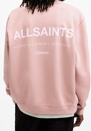 Osoba z ciemnymi dredami, ubrana w różową bluzę z napisem "ALLSAINTS ACCESS ALL AREAS BACKSTAGE LONDON" na plecach.