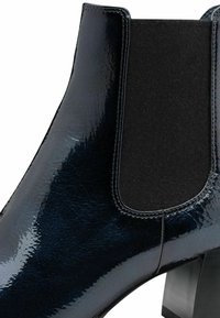 Bottine noire brillante avec panneau latéral élastique et petit talon bloc, montrant une surface en cuir lisse et texturée ainsi qu'une couture détaillée.
