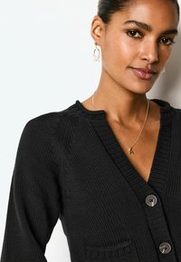 Sort strikket cardigan med V-udskæring, ribbet tekstur, to knapper og en lille lomme. Gyldne hoops og en delikat gylden halskæde fuldender looket.