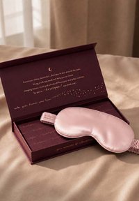 Roze zijden oogmasker dat rust op een open donkerpaarse doos met gouden tekst over luxe huidverzorging en ontspanning.