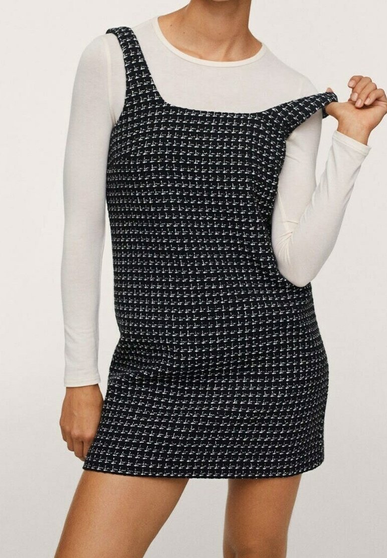 Femme portant une robe sans manches à carreaux noir et blanc par-dessus une chemise blanche à manches longues, tirant une bretelle vers l'extérieur.