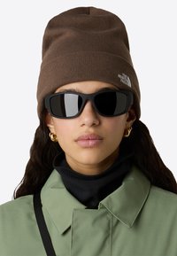 Osoba v hnědé pletené čepici s logem The North Face, černých obdélníkových slunečních brýlích, zlatých kruhových náušnicích, zelené bundě a černém roláku.