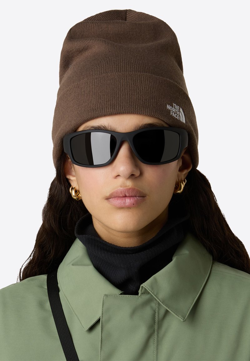 Osoba v hnědé pletené čepici s logem The North Face, černých obdélníkových slunečních brýlích, zlatých kruhových náušnicích, zelené bundě a černém roláku.