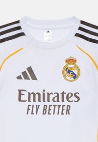 Weißes Sporttrikot mit schwarzen und goldenen Akzenten, das das Adidas-Logo, das Wappen von Real Madrid und den Text "Emirates Fly Better" auf der Vorderseite trägt.