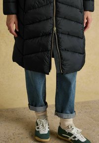 Melnā pūkainā jaka ar chevron piešuvumiem, zelta rāvējslēdzēju un sarullētām piedurknēm; zili džinsi; zaļi un balti sporta apavi; krēmkrāsas zeķes.