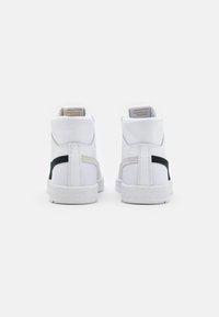 Paire de baskets hautes blanches avec des rayures latérales noires et grises, vues de dos sur un fond uni.