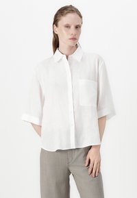 White linen short-sleeve shirt with a collar, button-front, and a single chest pocket. Features a loose fit and slightly sheer texture.