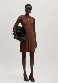 Bruine geribbelde jurk met knoopdetails bij de halslijn. Een zwarte glanzende tas en bruin gespenhakken maken de look compleet. Soepele textuur en aansluitende silhouet.