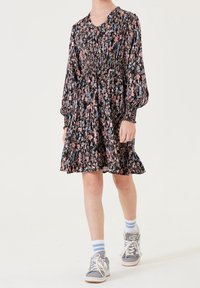 Czarna sukienka w kwiaty z bufkami na rękawach, talią zebrana i rozkloszowanym dołem. Noszona z szarymi trampkami i niebieskimi paskowanymi skarpetkami.