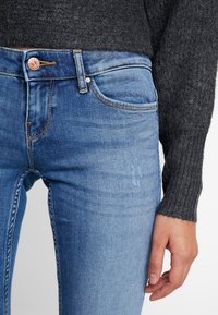 Vaqueros ajustados de color azul con un diseño desgastado, que presentan un botón de cobre, costuras naranjas y una parte de una camiseta de punto de manga larga gris oscuro visible.