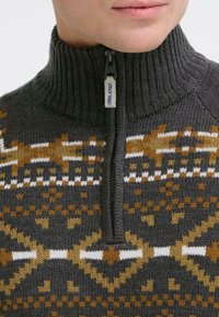 Mørkegrå strikket pullover med høj ribbet krave og lynlås. Har geometriske mønstre i brune, hvide og lysebrune nuancer på brystet.