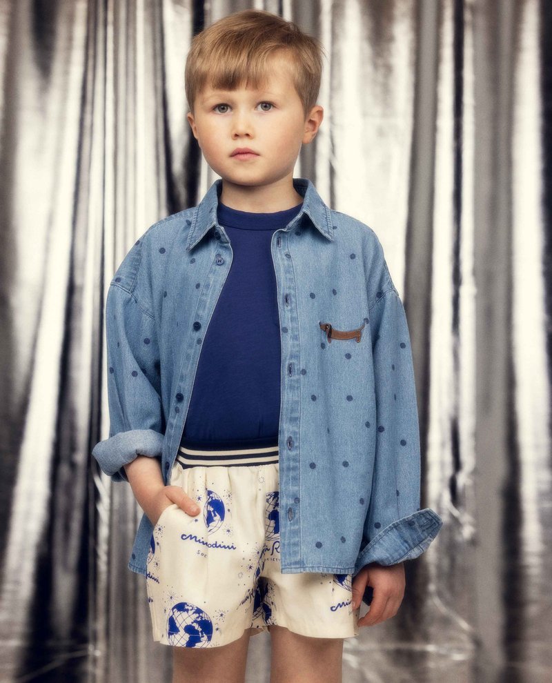 Νεαρός αγόρι με ανοιχτό καστανό μαλλί φοράει μπλε πουά τζιν πουκάμισο πάνω από μπλε ναυτικό μπλουζάκι και κρεμ σορτς με μπλε εκτυπώσεις παγκόσμιου χάρτη και κείμενο.