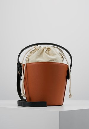 Bruine leren buideltas met zwart handvat en riem, beige stoffen zakje met trekkoord binnenin, geplaatst op een witte ondergrond.