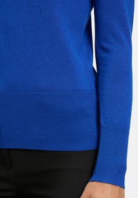 Blauer Strickpullover mit gerippten Bündchen und Saum; glatte Textur; figurbetontes Design, kombiniert mit schwarzem Stoff darunter.