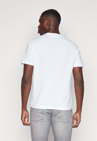 Hvit t-skjorte med korte ermer laget av bomull, med rund hals og avslappet passform. Brukt med grå jeans, vist fra bak.