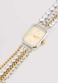 Ceas metalic aurie și argintie, cu un cadran rectangular, un dial galben și accente cu cristale; dispune de o brățară multi-link cu lanțuri și pietre prețioase.