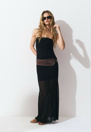 Black maxi dress with a fitted bodice, sheer pleated skirt, and brown belted accent. Model wears dark sunglasses and flat sandals.