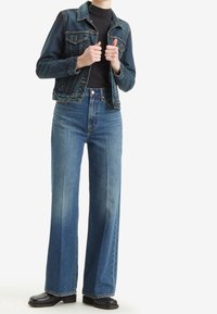 Personne portant une veste en jean bleu foncé, un haut noir, un jean bleu taille haute à jambes larges et des chaussures noires, debout les mains ajustant l'avant de la veste.