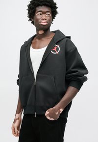 Μαύρο φούτερ με φερμουάρ και τσέπες μπροστά, με κόκκινο και ασημένιο κυκλικό λογότυπο. Απαλή, λεία ύφανση με άνετη εφαρμογή και κουκούλα.