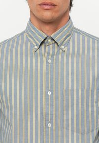 Camicia azzurro chiaro con righe verticali gialle, colletto e una tasca sul petto, con bottoni bianchi e texture in cotone liscio.