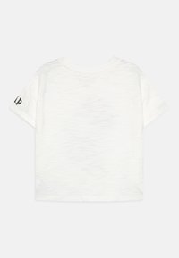 Balta īsām piedurknēm T-kreklā ar strukturētu, nedaudz caurspīdīgu audumu. Tam ir apaļa kakla izgriezums un mazs melns logotips kreisajā piedurknē.