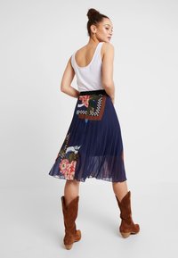 Gonna plissettata blu navy con motivo floreale e toppe a contrasto. Il modello indossa una canotta bianca e stivali alla caviglia in suede marrone.